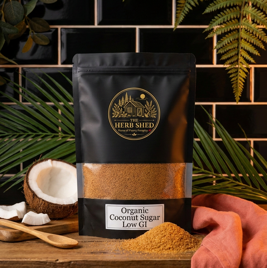 A black stand-up pouch of Herb Shed Organic Coconut Sugar Low GI sits on a wooden surface with a coconut half, wooden spoon, and loose sugar in front. The pouch has a clear window showing the brown sugar inside.