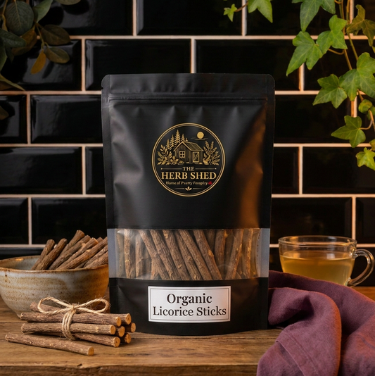 A black stand-up pouch of Herb Shed Organic Licorice Sticks sits on a wooden table against a black tiled wall, with a bowl of sticks, a glass of tea, and greenery around it.
