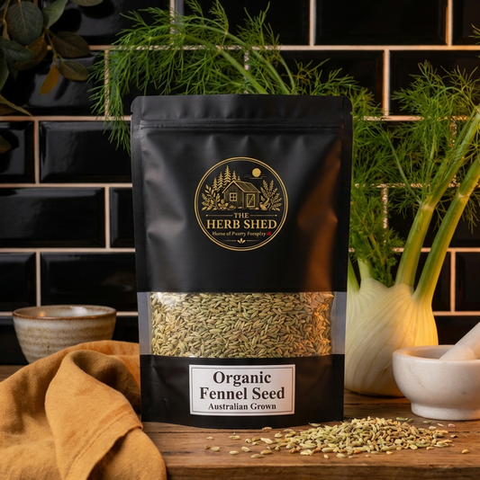 A black stand-up pouch of Herb Shed Organic Fennel Seed displayed on a wooden surface with a black tiled background, flanked by fresh fennel bulbs, a mustard cloth, and small bowls.