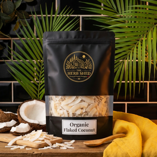 A black stand-up pouch of Herb Shed Organic Flaked Coconut stands on a wooden surface with coconut halves, shreds, and a yellow cloth. The background features a black tiled wall and green palm leaves.