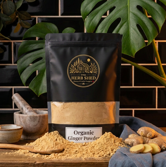 A black stand-up pouch of Herb Shed Organic Ginger Powder sits on a wooden surface with loose ginger powder spilled in front, ginger roots, a blue cloth, and large green monstera leaves against a dark tiled background.