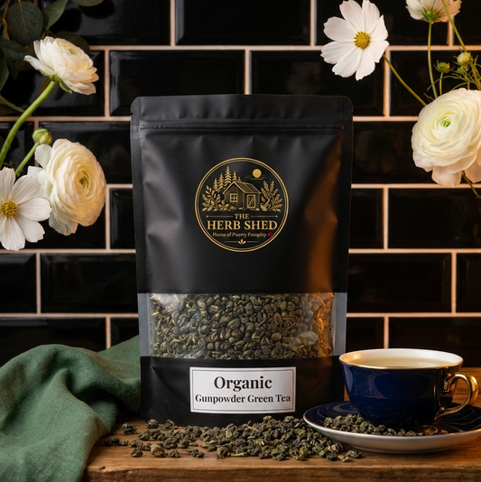 A black stand-up pouch of Herb Shed Organic Camomile Green Tea sits on a wooden surface against a black tiled wall, with white flowers draped around it and a blue teacup with tea beside the package.