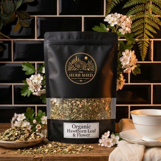 A black stand-up pouch of Herb Shed Organic Hawthorn Leaf & Flower tea, displayed on a rustic wooden table with a black tiled background, green ferns, white blossoms, and a white teacup.