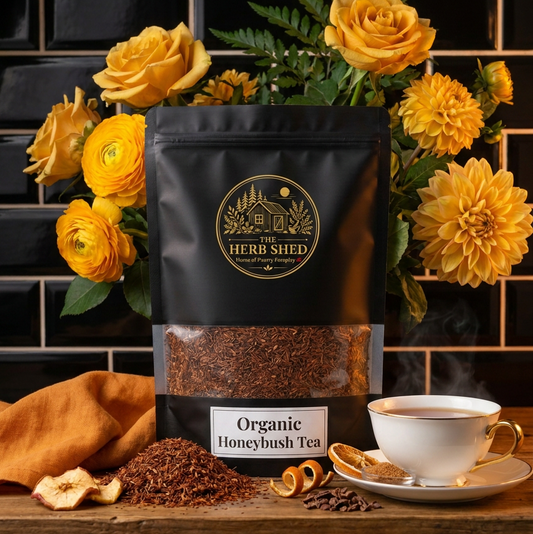 A black stand-up pouch of Herb Shed Organic Honeybush Tea stands on a wooden surface with loose tea, dried fruit, citrus slices, and a white teacup against a black tiled wall and yellow flowers.