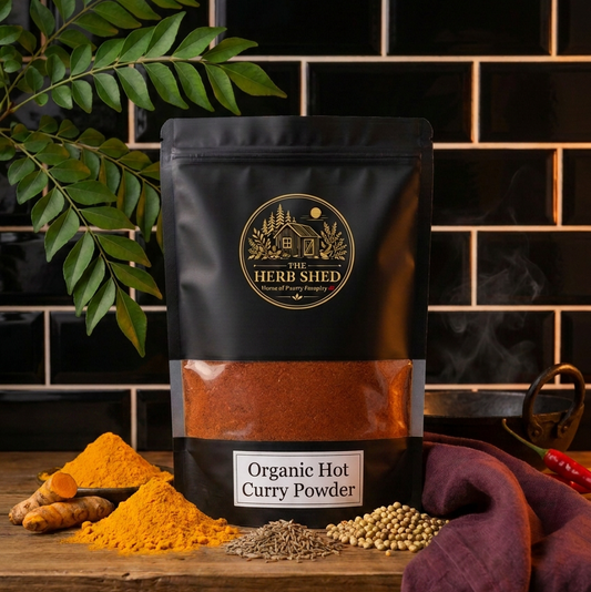 A black stand-up pouch of Herbi Shed Organic Hot Curry Powder sits on a wooden surface with curry powder, turmeric root, and spices scattered around it. The background features a black subway tile wall with a green leafy branch on the left.
