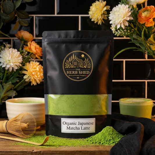 A black stand-up pouch of Herb Shed Organic Japanese Matcha Latte sits on a wooden surface with green matcha powder spilled in front. The pouch has a clear window showing bright green matcha inside, with a white label reading the product name.