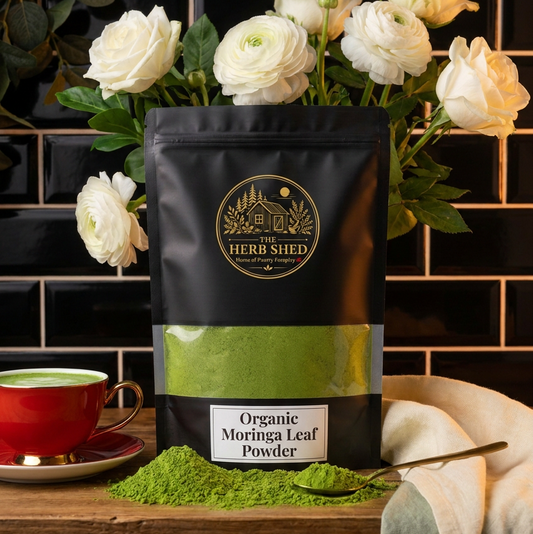 A black stand-up pouch of Herbs Shed Organic Moringa Leaf Powder sits on a wooden surface with green powder spilled in front, a spoon, a red cup of green tea, and white flowers behind it against a black tiled wall.