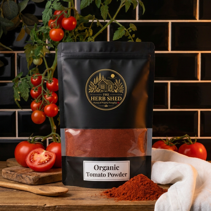 A black stand-up pouch of Herb Shed Organic Tomato Powder stands on a wooden surface with loose red tomato powder spilled in front. Fresh tomatoes, a cutting board, and a white cloth are arranged around it, with a black tiled wall in the background.