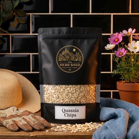 A black stand-up pouch of Herb Shed Quassia Chips sits on a wooden surface with scattered chips in front. The pouch has a clear window showing the light tan chips inside and a white label reading 'Quassia Chips.
