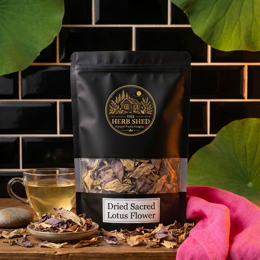 A black stand-up pouch of Heri Shod Dried Sacred Lotus Flower tea stands on a wooden surface against a black tiled wall, with dried flower pieces scattered around, a glass of brewed tea, and a pink cloth nearby. Large green lotus leaves frame the top of the scene.