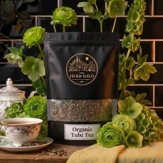 A black stand-up pouch of Herb Shed Organic Tulsi Tea is displayed on a wooden surface, surrounded by fresh green tulsi leaves and flowers, with a teacup and dark tiled background.