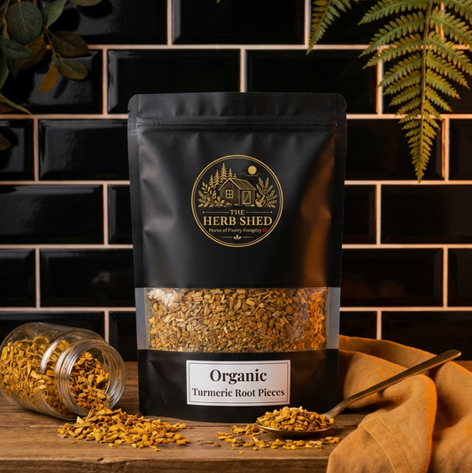 A stand-up pouch of Herb Shed Organic Turmeric Root Pieces sits on a wooden surface against a black subway tile wall, with loose turmeric pieces spilled from a glass jar and a spoonful nearby. Fern leaves and a mustard cloth frame the scene.