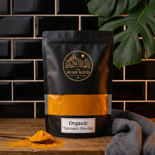 A black stand-up pouch of Heri Shid Organic Turmeric Powder with a gold logo and a horizontal orange band, displayed on a wooden surface with a small spoonful of yellow powder in front, against a black tiled wall with a green monstera leaf.