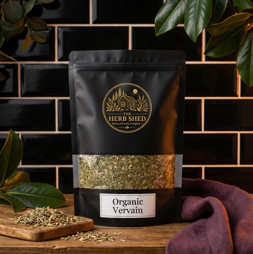A black stand-up pouch of Herb Shed Organic Vervain tea or herb product, displayed on a wooden surface against a black subway tile wall with green leaves and a purple cloth around it.