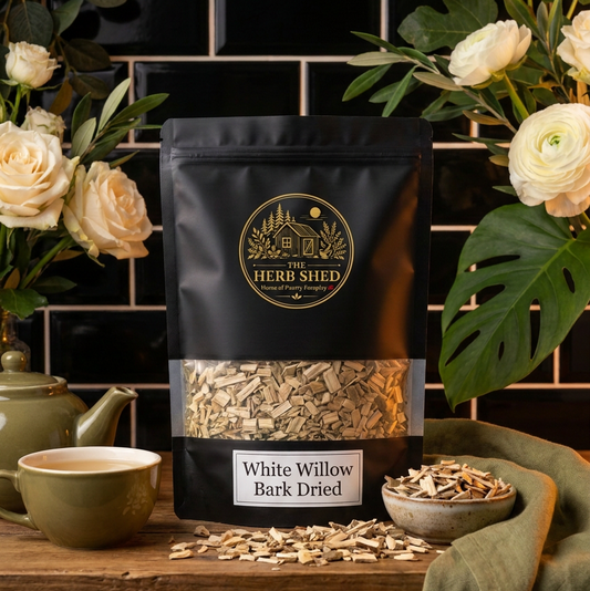 A black stand-up pouch of Herb Shed White Willow Bark Dried, styled on a rustic wooden table with a green ceramic teapot, cup, bowl of bark pieces, and white roses against a black subway tile background.