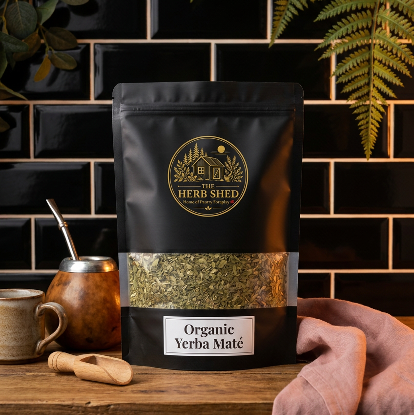 A black stand-up pouch of Herb Shed Organic Yerba Maté tea with a clear window showing the dried green leaves inside, styled on a wooden surface with a mate cup, wooden scoop, and pink cloth against a black tiled background.
