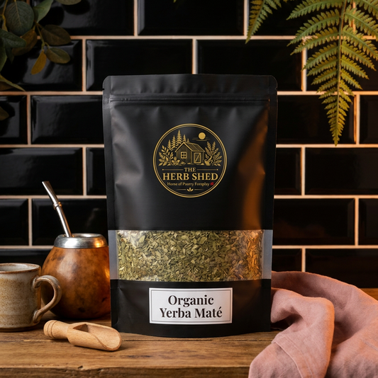 A black stand-up pouch of Herb Shed Organic Yerba Maté tea with a clear window showing the dried green leaves inside, styled on a wooden surface with a mate cup, wooden scoop, and pink cloth against a black tiled background.