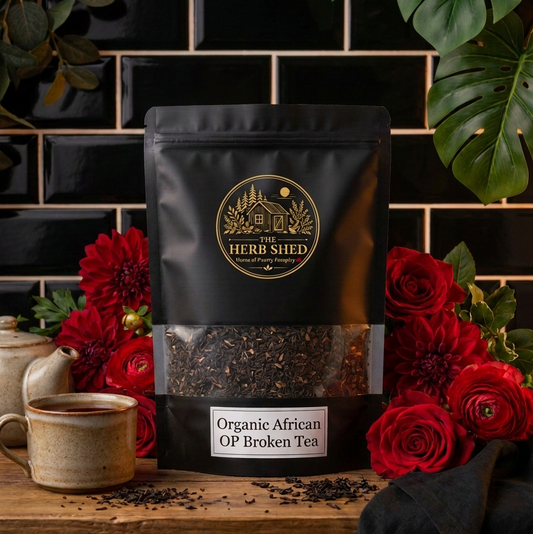 A black stand-up pouch of Heri Shid Organic African OP Broken Tea sits on a wooden surface, surrounded by red flowers, green leaves, a ceramic mug, and a dark tiled background.
