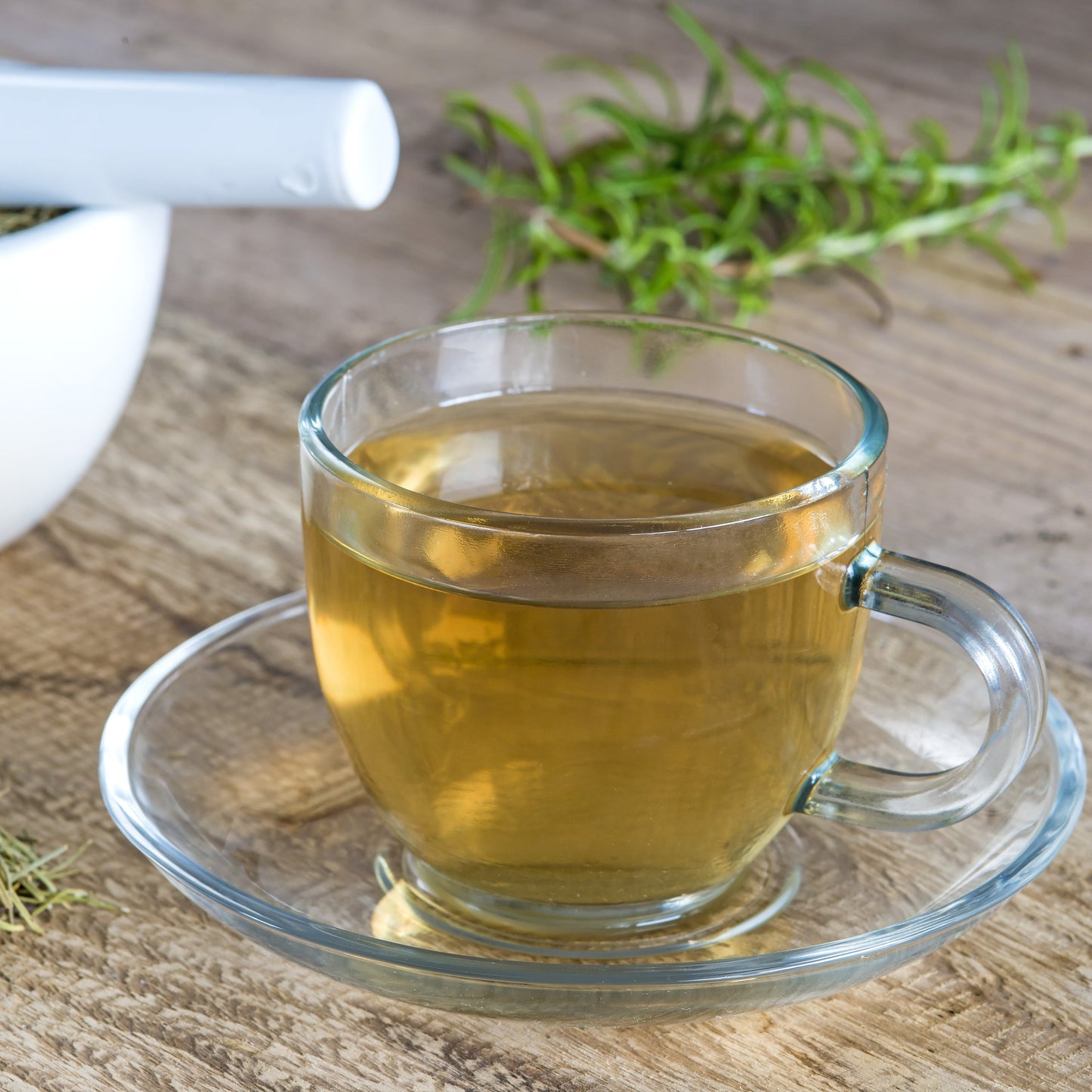A clear glass cup filled with amber tea on a matching saucer, set on a rustic wooden table beside a white mortar and pestle containing dried herbs, with loose rosemary sprigs nearby.