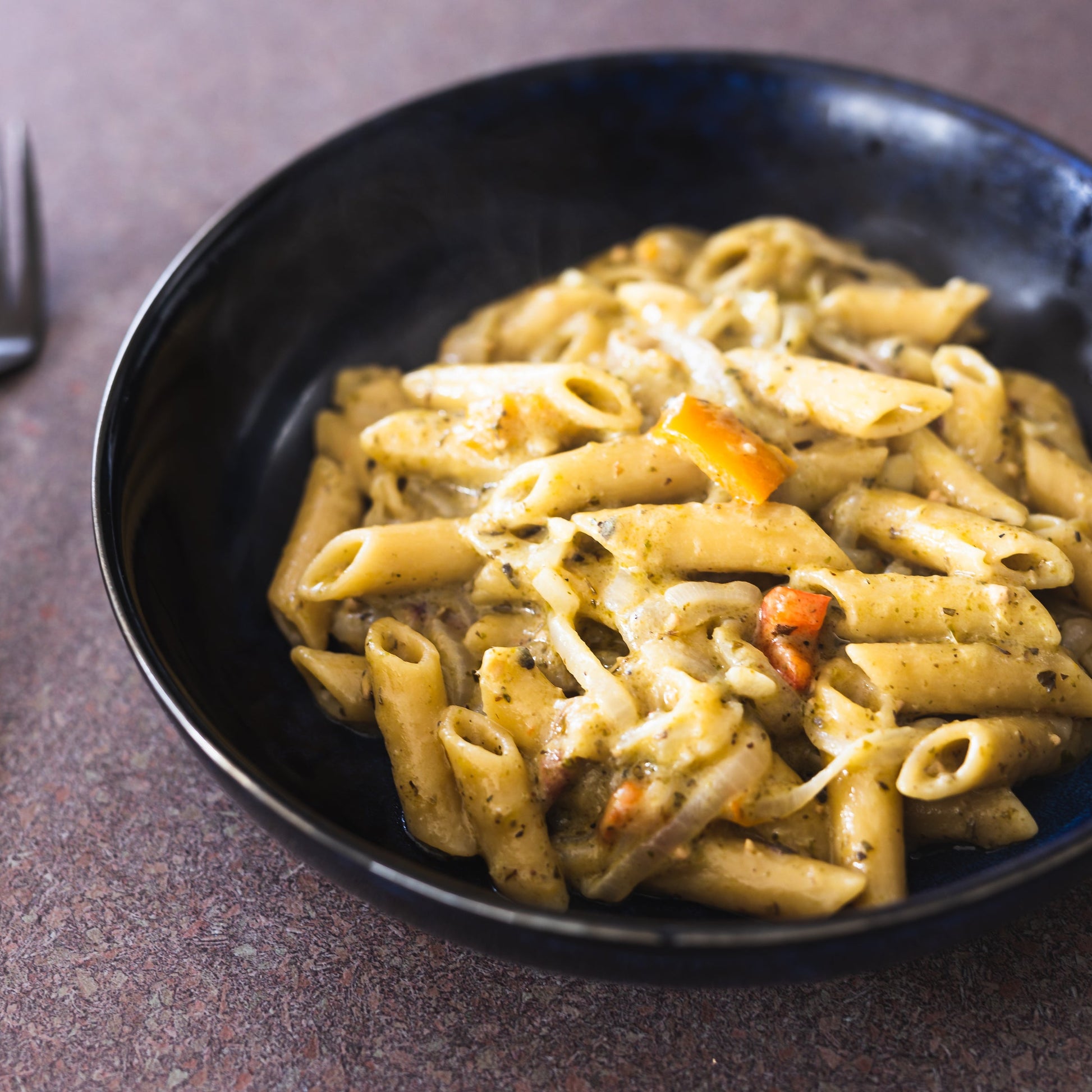 A bowl of penne pasta with a creamy mushroom and vegetable sauce, served in a dark blue ceramic bowl with a black fork on a textured gray surface.