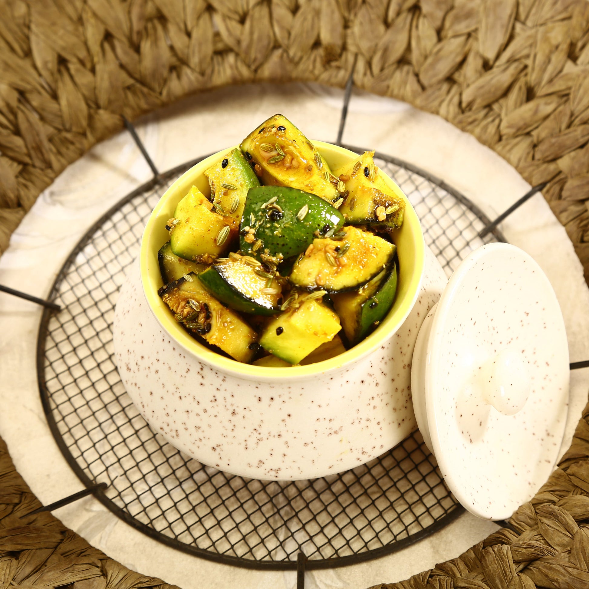 A bowl of cooked diced zucchini and yellow squash with herbs, served on a small speckled ceramic dish with a lid, set on a black wire cooling rack over a woven brown placemat.