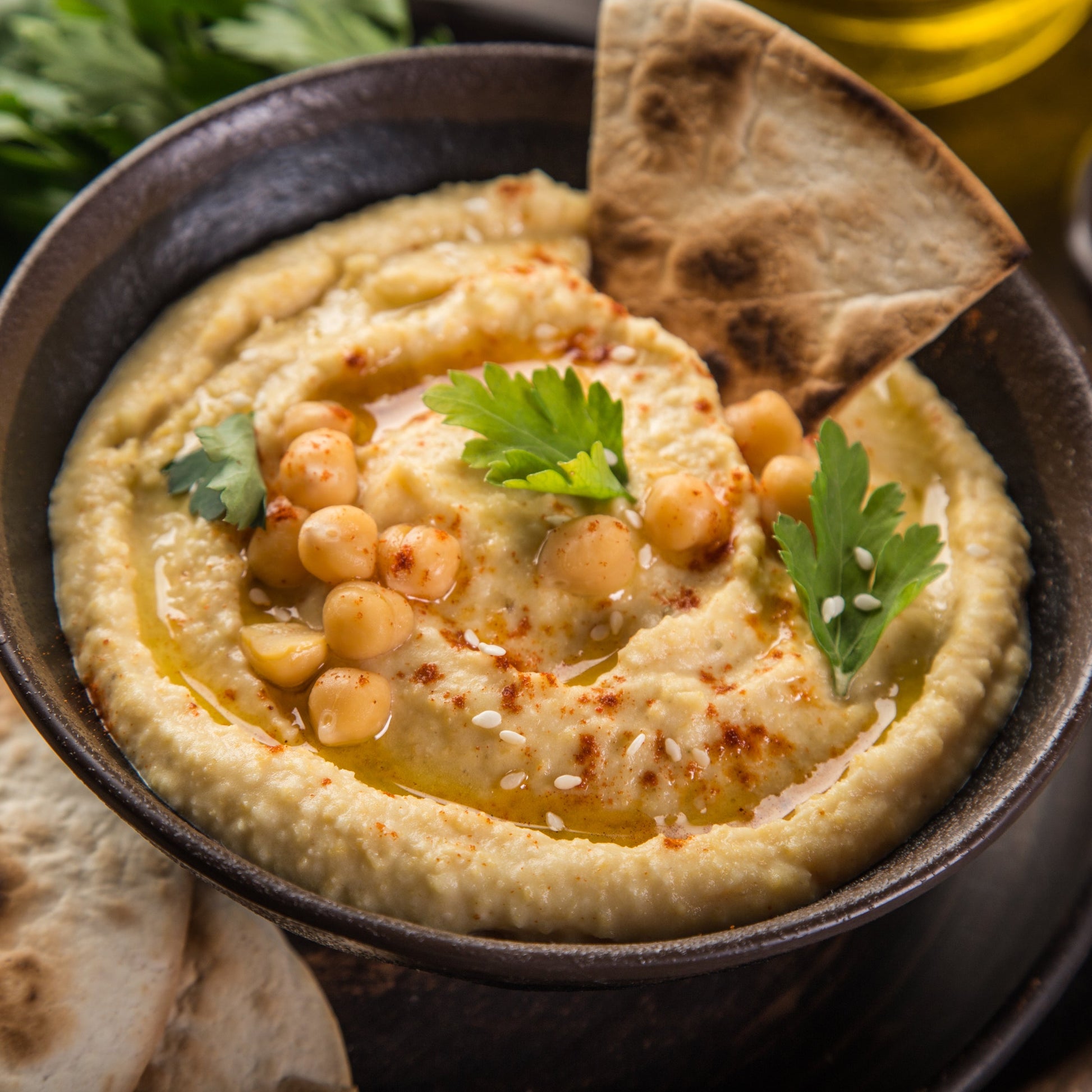 A close-up of a bowl of chickpea hummus topped with olive oil, parsley, and smoked paprika, served with pita bread on a dark rustic background.