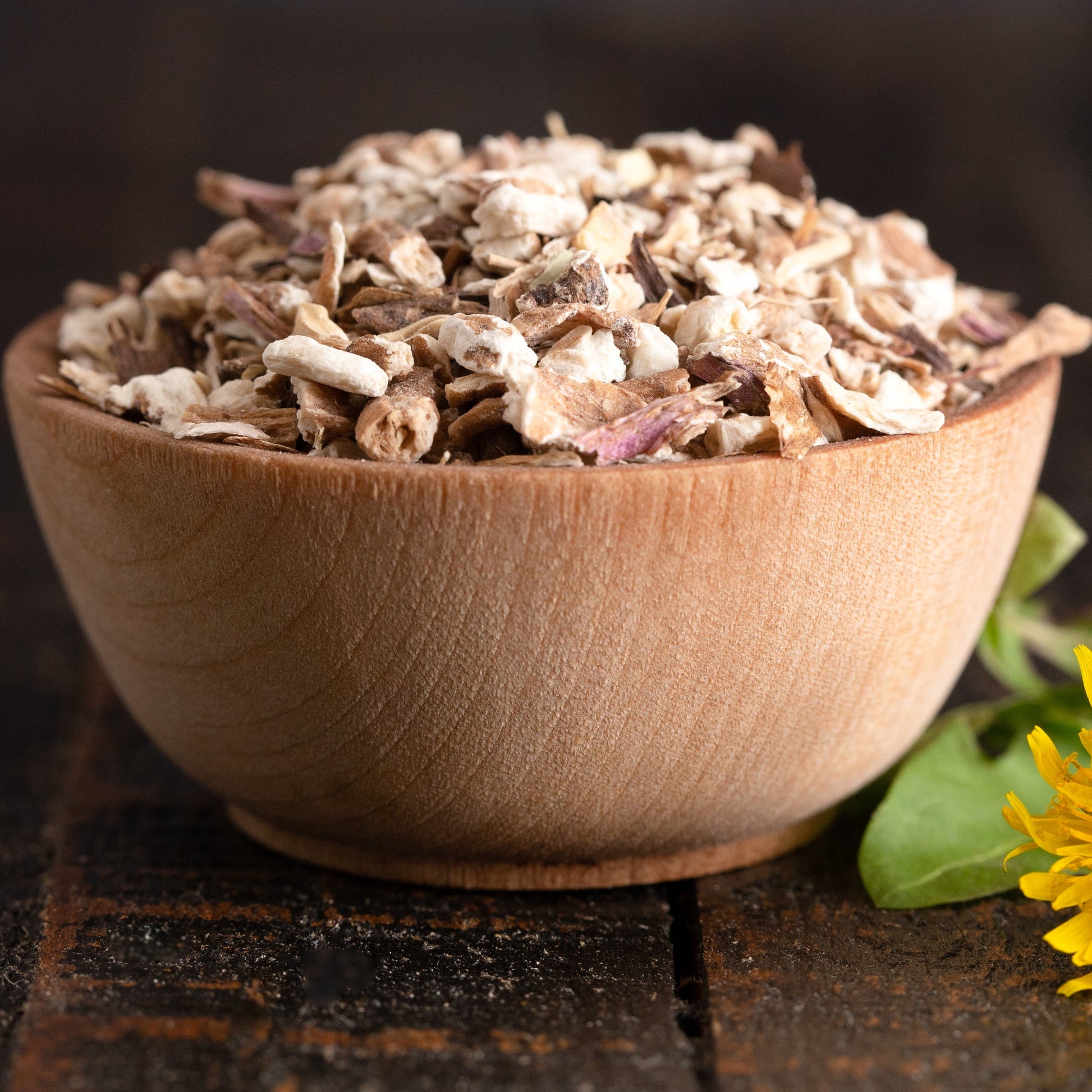 A small wooden bowl filled with dried oats and herbs, set on a dark rustic wooden surface next to a yellow dandelion flower and green leaves.