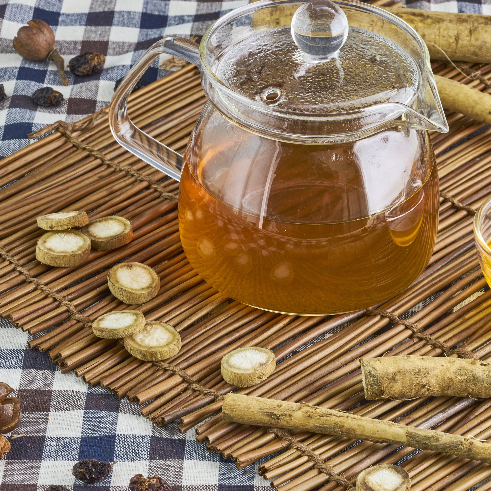 A clear glass teapot filled with amber tea sits on a woven bamboo mat beside a small glass cup of golden liquid, surrounded by sliced and whole herbal roots on a blue-and-white checkered cloth.