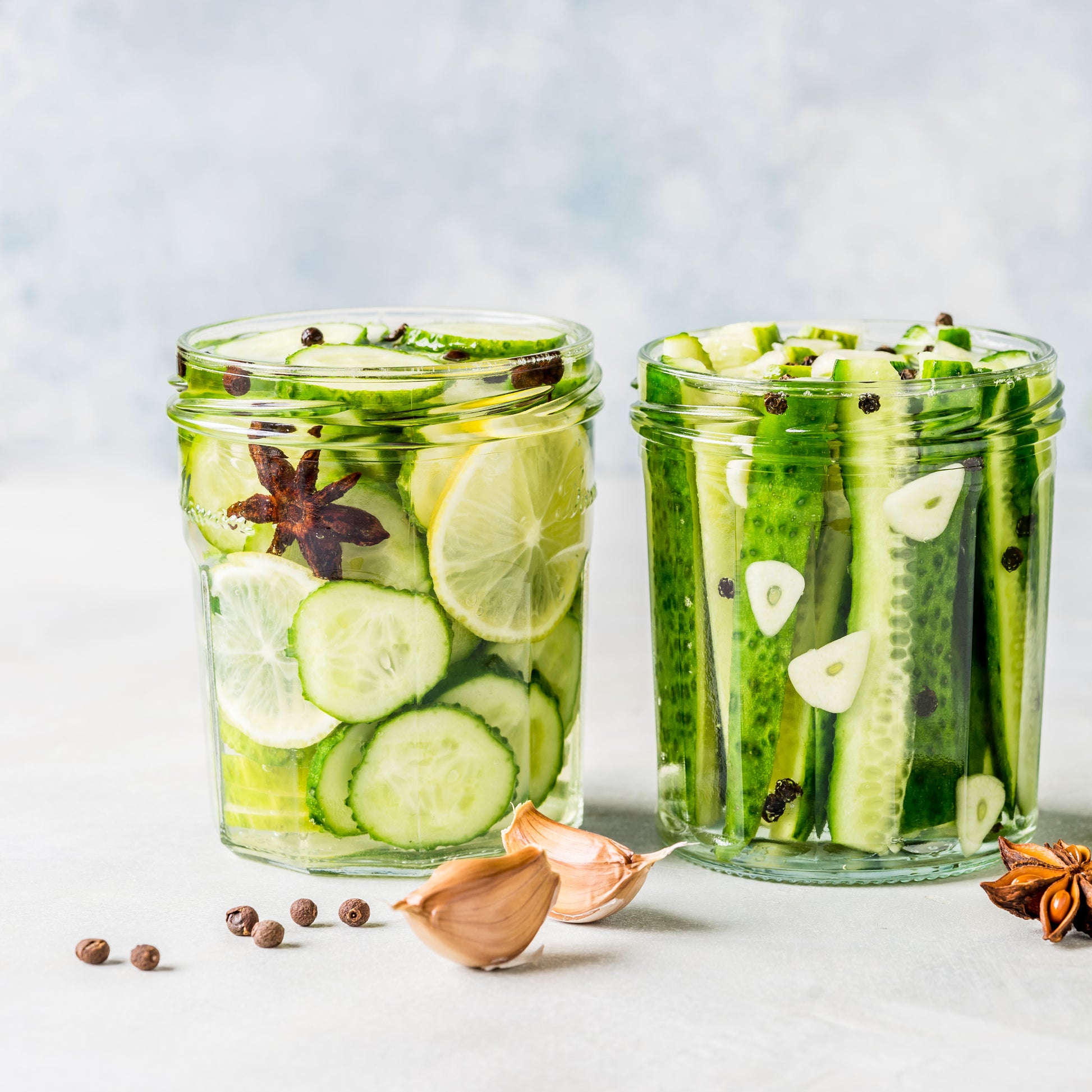 Two clear glass jars filled with pickled cucumbers and lime slices, styled on a light surface with garlic cloves, peppercorns, and star anise.
