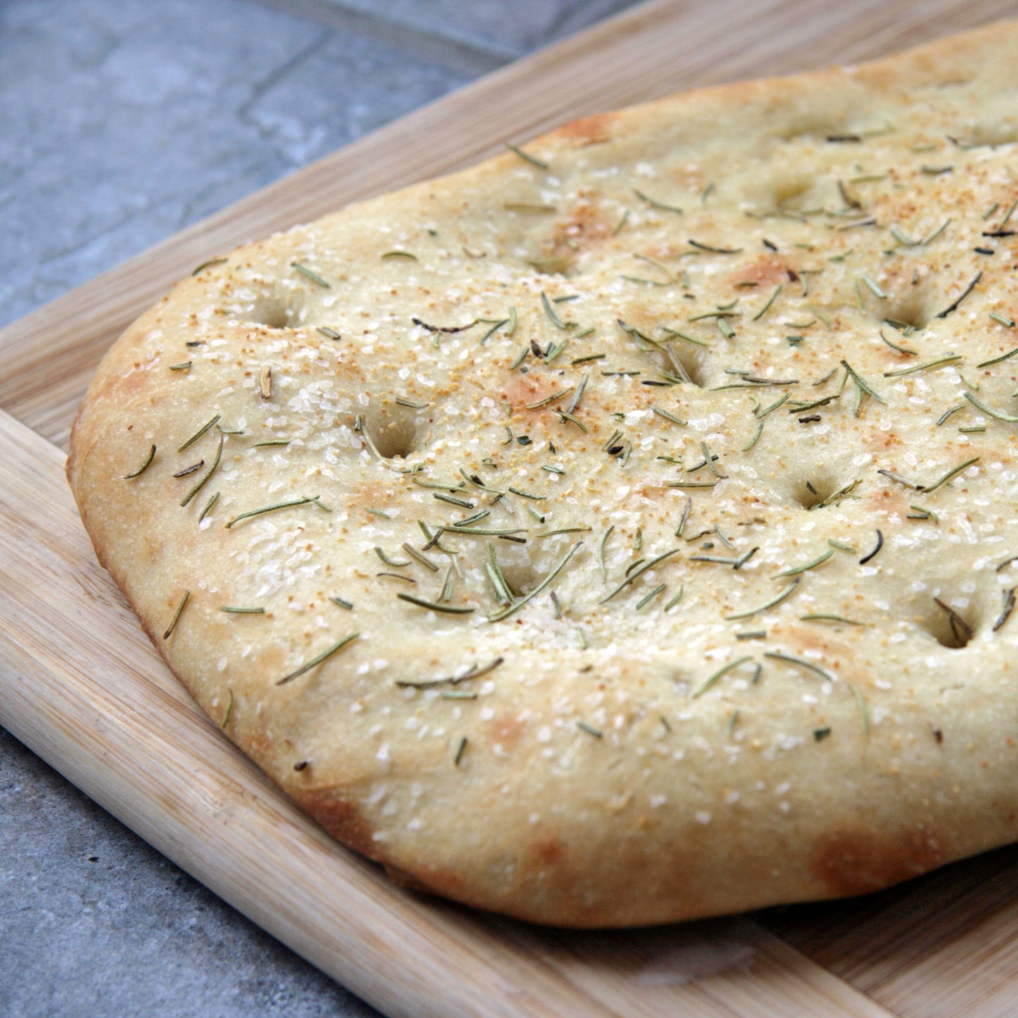 A freshly baked square focaccia bread topped with herbs and coarse sea salt, resting on a wooden cutting board.