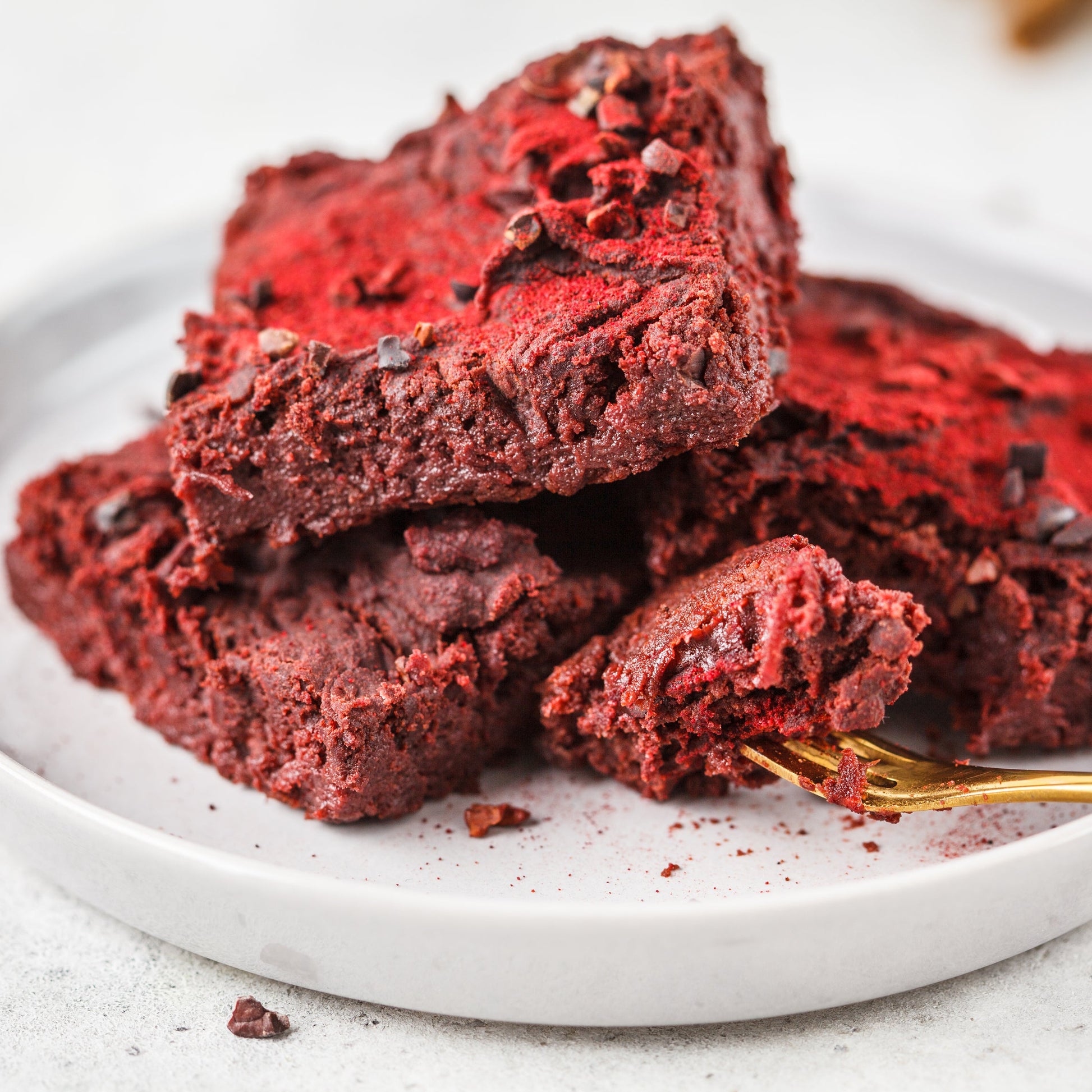 Rich chocolate beetroot brownies are stacked on a white plate with red crumbs scattered around them, showing a baking idea using beetroot powder.