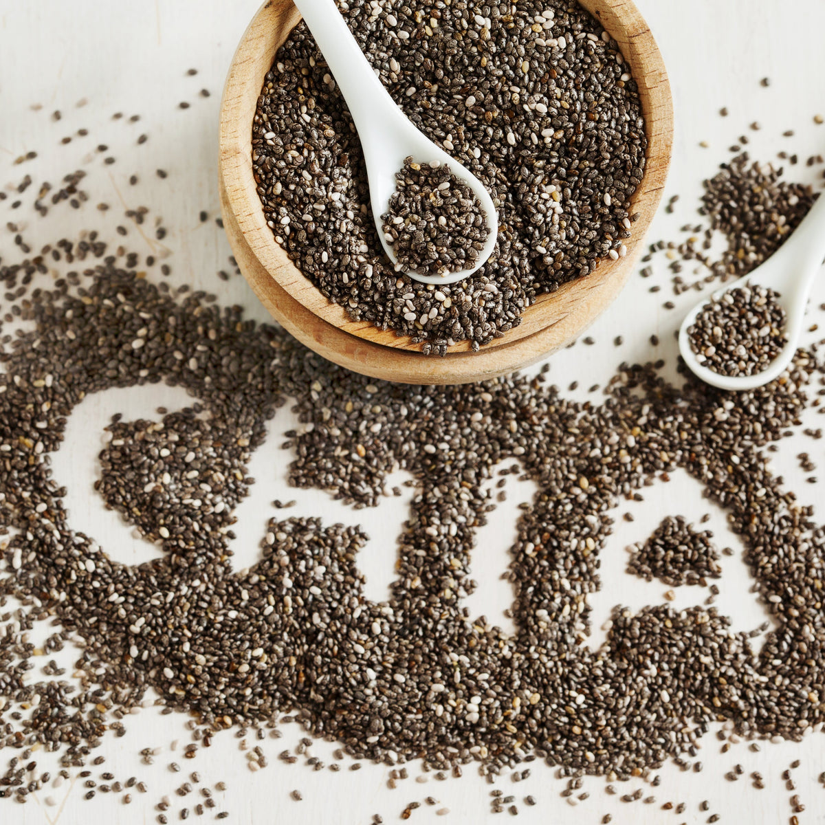 Black Chia Seeds spill from a wooden scoop onto a white surface, showing the small mottled seeds and their natural texture.