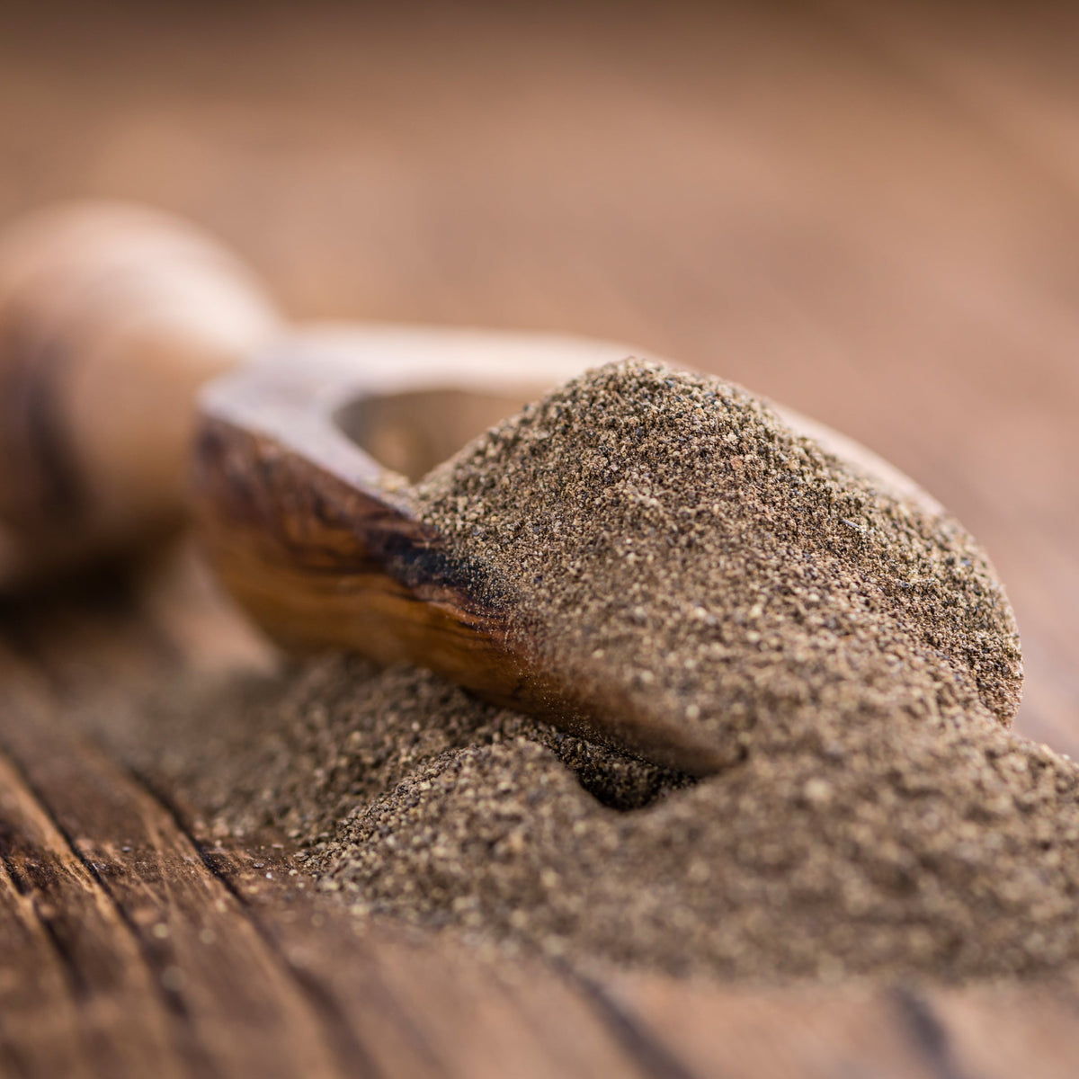 Ground black pepper fills a small wooden scoop on a rustic wooden surface, showing its finely ground texture and natural dark speckled colouring.