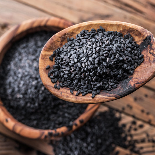 Black Sesame Seeds fill a wooden spoon with loose seeds scattered around it on a rustic wooden surface, showing their small size and natural black colouring.