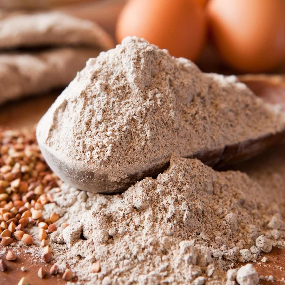 A mound of Buckwheat Flour on a rustic surface beside eggs, showing the flour's finely milled texture and natural light brown colouring.