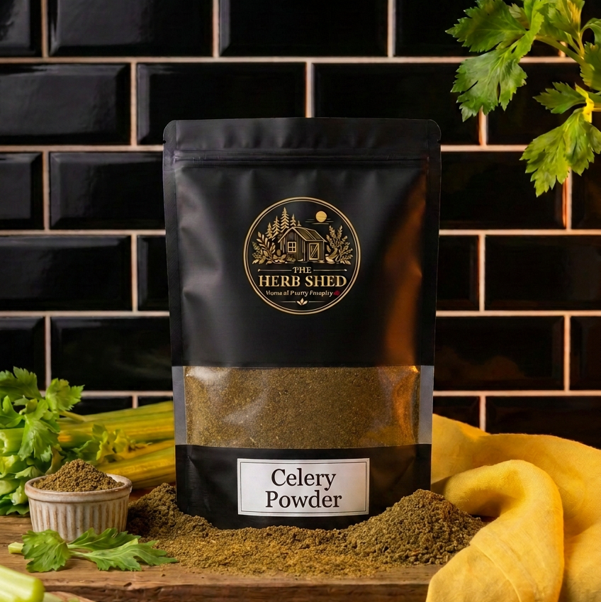 A black stand-up pouch of The Herb Shed Celery Powder sits on a wooden surface with loose pale green celery powder, fresh celery leaves and a small bowl, with soft yellow fabric against a black tiled background.