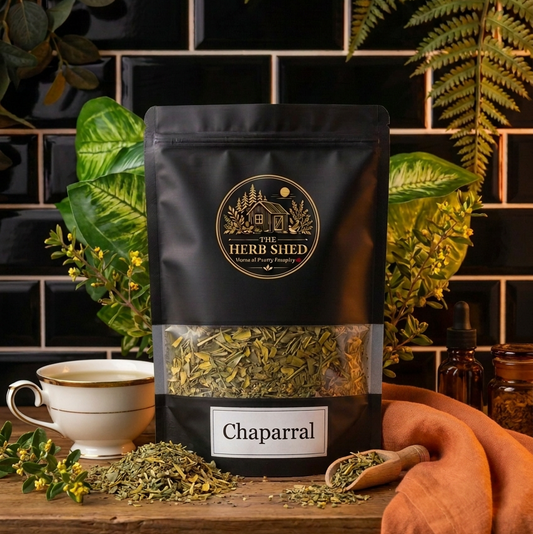 A black stand-up pouch of The Herb Shed Chaparral sits on a wooden surface with loose dried chaparral, a wooden scoop and a cup of herbal tea, surrounded by leafy greenery and soft rust-coloured fabric against a black tiled background.