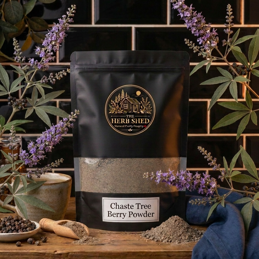 A black stand-up pouch of The Herb Shed Chaste Tree Berry Powder sits on a wooden surface with loose berry powder, whole dried chaste tree berries, a wooden scoop and purple flowering branches, against a black tiled background with deep blue fabric.