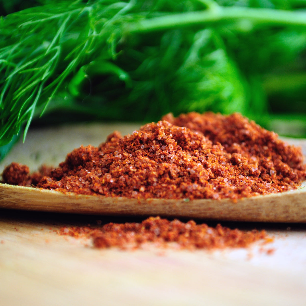 Chermoula Spice fills a wooden spoon beside fresh green herbs, showing the seasoning's coarse texture and warm red-orange colouring with visible spice flecks.