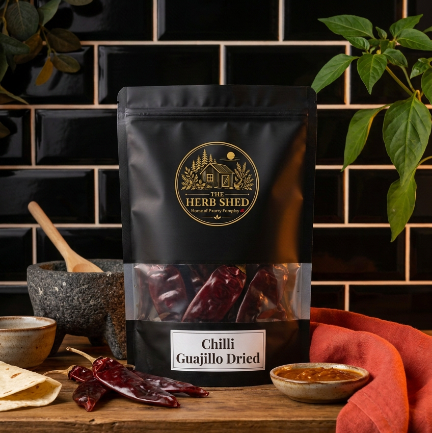 A black stand-up pouch of The Herb Shed Chilli Guajillo Dried sits on a wooden surface with whole dried guajillo chillies, a mortar and pestle, tortillas and a small bowl of sauce, surrounded by green foliage and warm red fabric against a black tiled background.