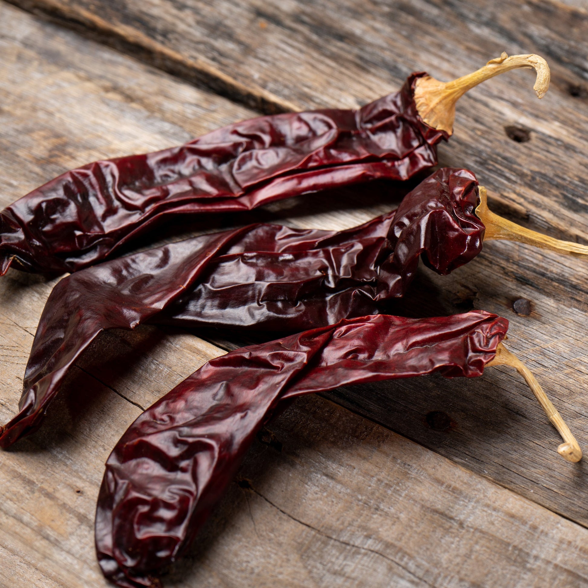 Whole dried guajillo chillies are arranged on a rustic wooden surface, showing their long shape, wrinkled skin and deep red-brown colour.