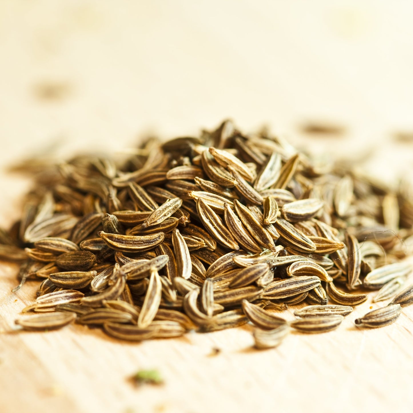 A small pile of whole cumin seeds sits on a light surface, showing their elongated shape, ridged texture and natural brown colouring.