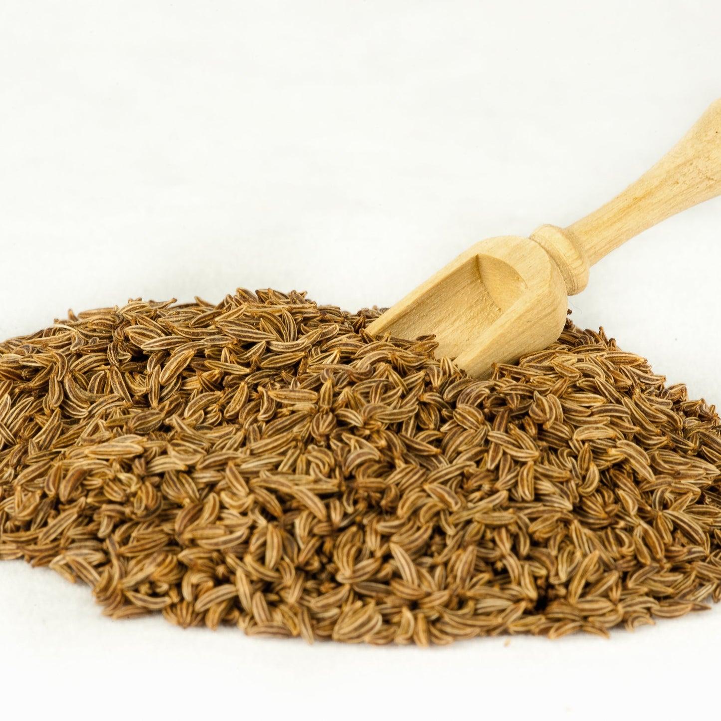 Whole cumin seeds are piled on a white surface beside a small wooden scoop, showing their slender shape and warm brown colour.