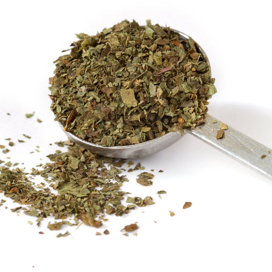 Dried basil flakes heaped in a metal measuring spoon with scattered herb pieces on a white surface, The Herb Shed, Wollongong