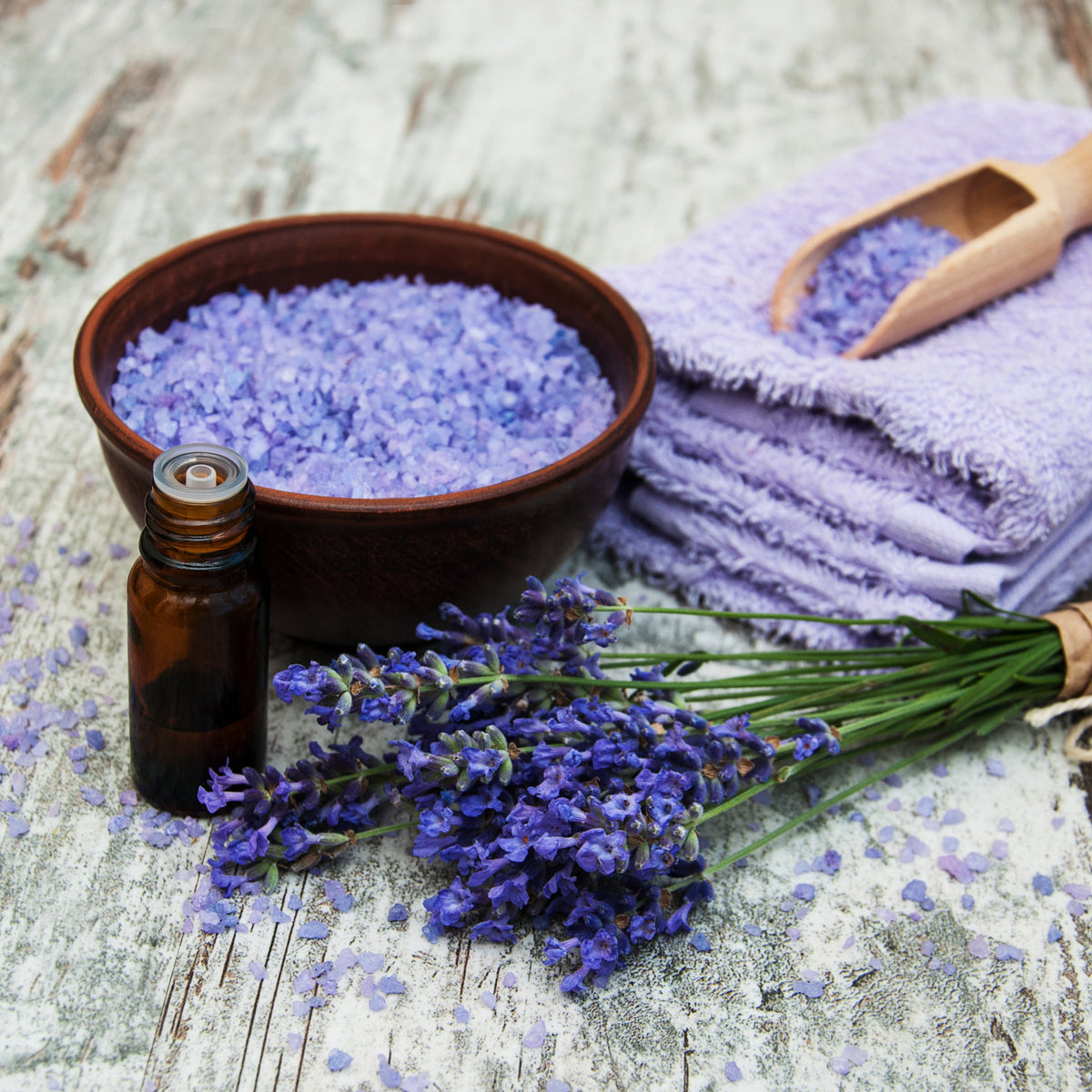 A bowl of purple bath salts beside dried lavender flowers and folded towels, showing Dried Lavender Flowers from The Herb Shed in Wollongong, Australia, used in a bath and body setting.