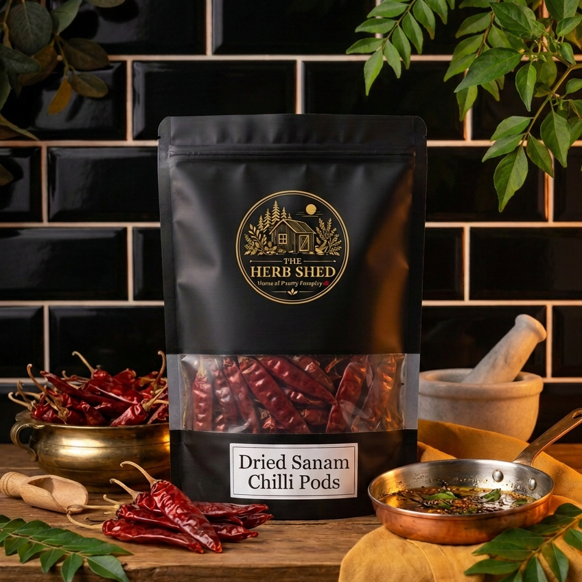 A black stand-up pouch of The Herb Shed Dried Sanam Chilli Pods sits on a wooden surface with whole dried red chillies, a brass bowl, mortar and pestle, fresh green foliage and a small bowl of chilli sauce, against a black tiled background.