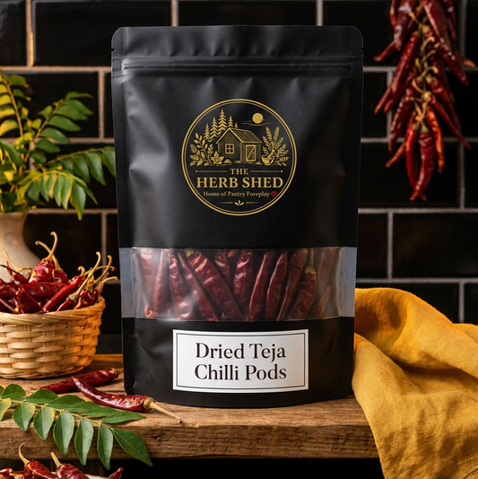 A black stand-up pouch of The Herb Shed Dried Teja Chilli Pods sits on a wooden surface surrounded by whole dried red chillies, green foliage, a woven basket and warm mustard-coloured fabric, against a black tiled background.