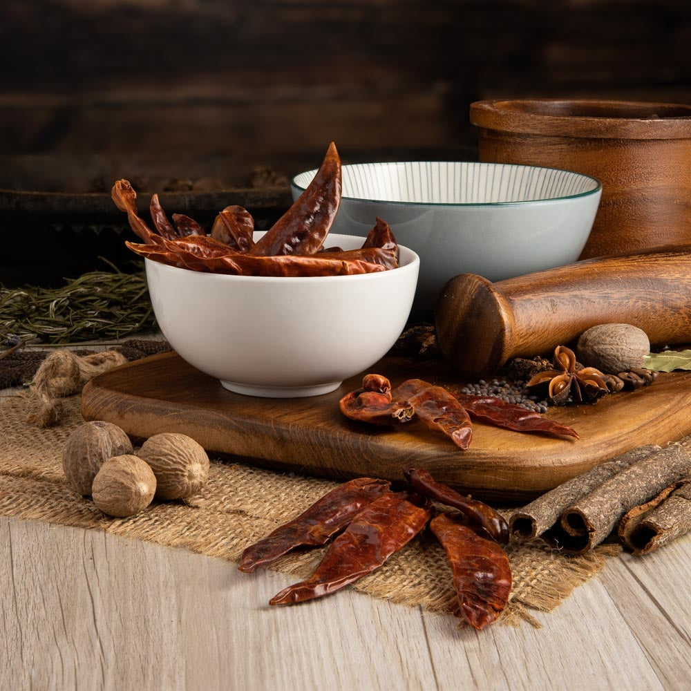 Whole dried red chillies are arranged with assorted whole spices and a small white bowl on a rustic wooden surface, showing a cooking use for dried Teja chilli pods.