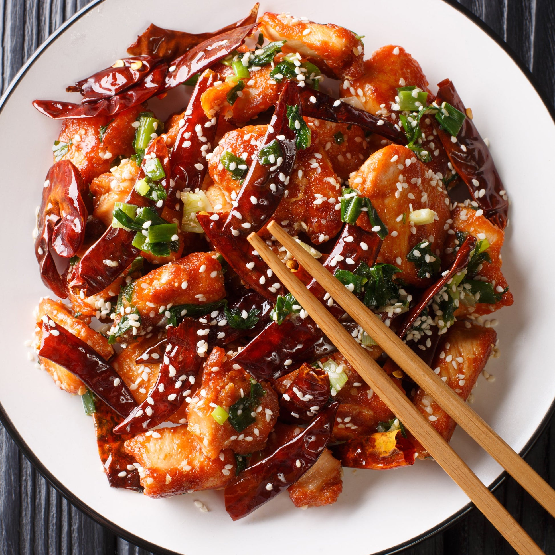 A spicy stir-fry made with dried red chillies is served on a white plate and topped with sesame seeds and green herbs, with chopsticks resting over the dish.