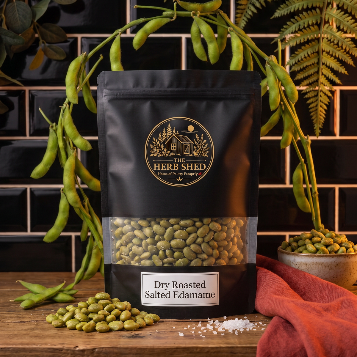 A black stand-up pouch of Dry Roasted Salted Edamame displayed with edamame and leafy foliage in a dark kitchen setting, from The Herb Shed Wollongong Australia.